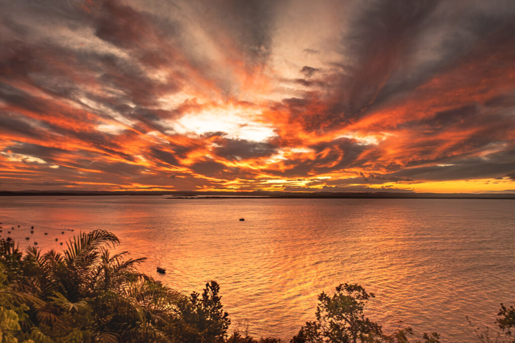 Céu dramático em tons de laranja e nuvens densas sobre o mar, com vegetação escura em primeiro plano.