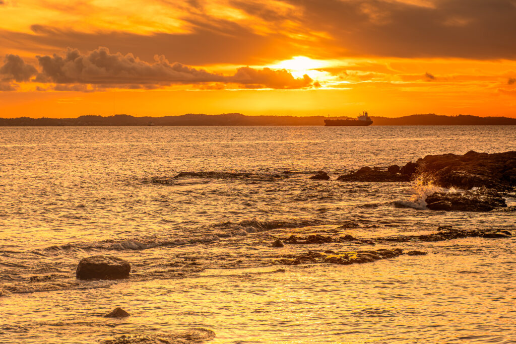 Pôr do sol sobre a baía, com um navio ao longe e ondas quebrando nas rochas da margem.