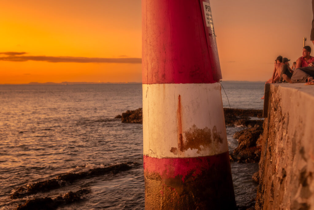 Farol de listras vermelhas e brancas diante do pôr do sol, junto ao mar.