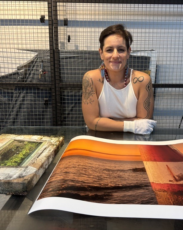 Alanna Sousa à mesa do ateliê, de luvas brancas, ao lado de obras impressas do DROP 01.